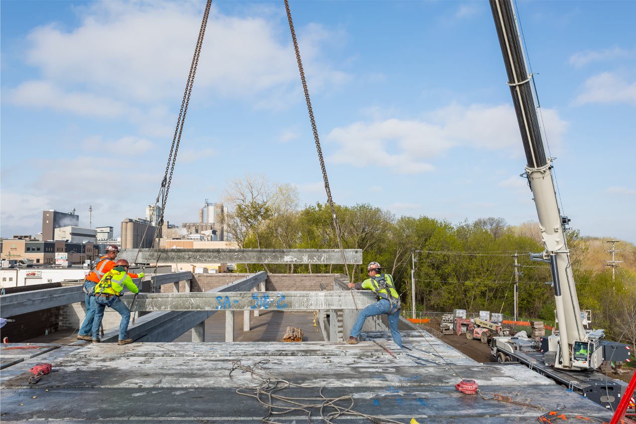 Three construction workers wearing hard hats and high-viz gear position a hollowcore plank on a podium with the help of a crane.