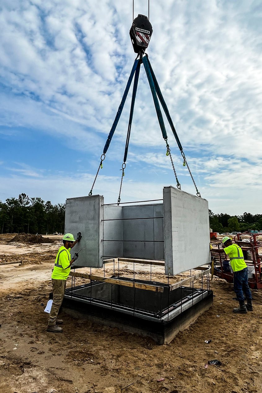 Construction workers guide a segment of a precast elevator shaft onto a foundation with the help of a crane.