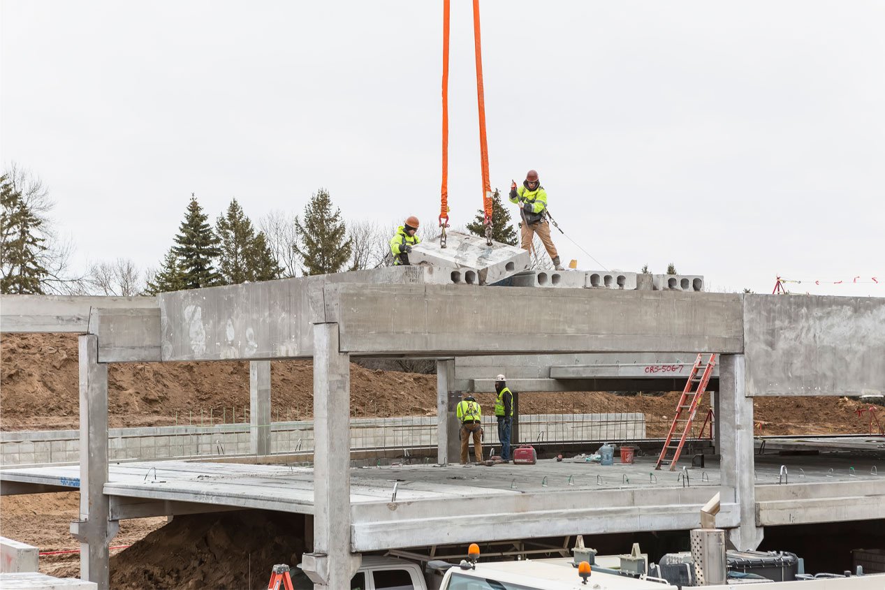 Construction workers position hollowcore plank into place on a construction site with the help of a crane.