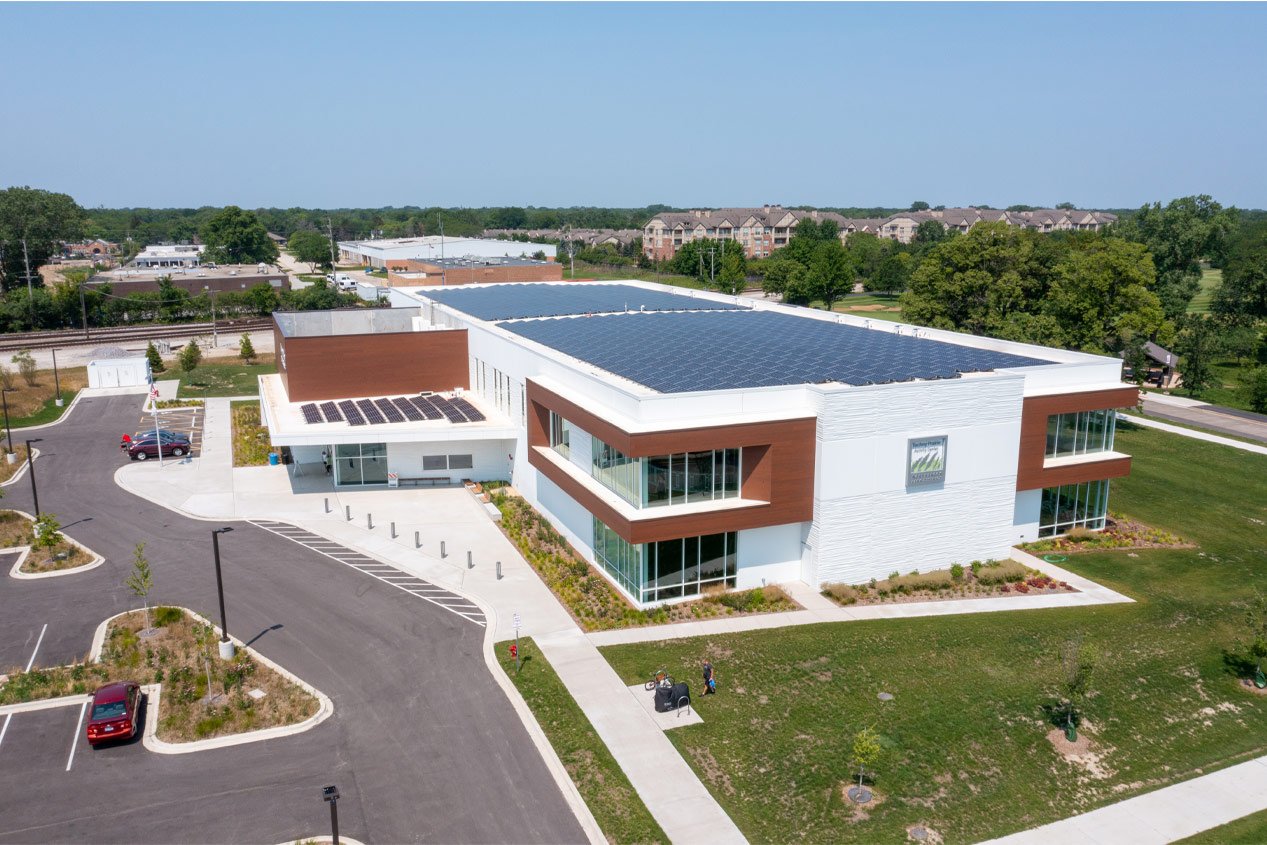 A two-story modern building with white precast concrete panels and brown accents and solar panels on the roof. 