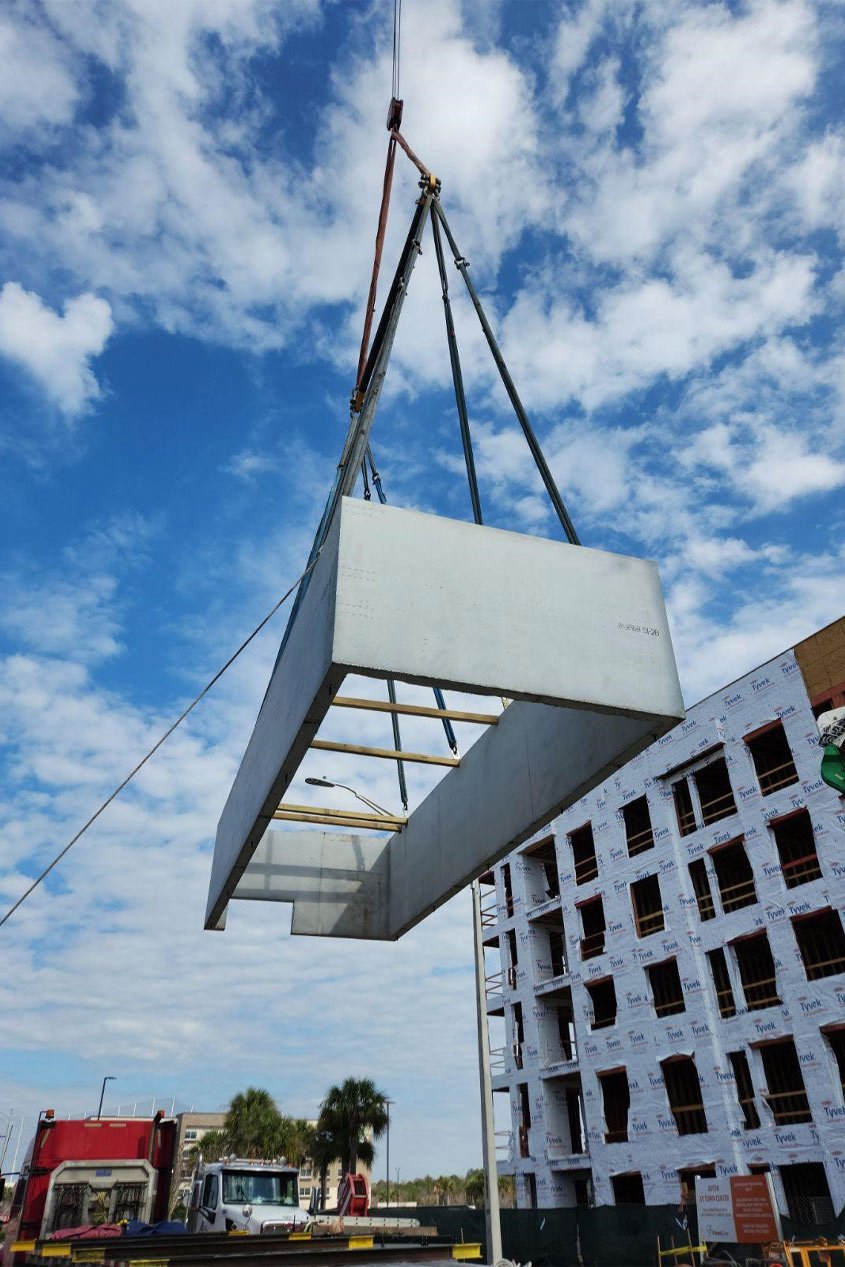One section of a precast concrete stair shaft is raised into the air by a crane in front of a multi-story building that is still under construction.