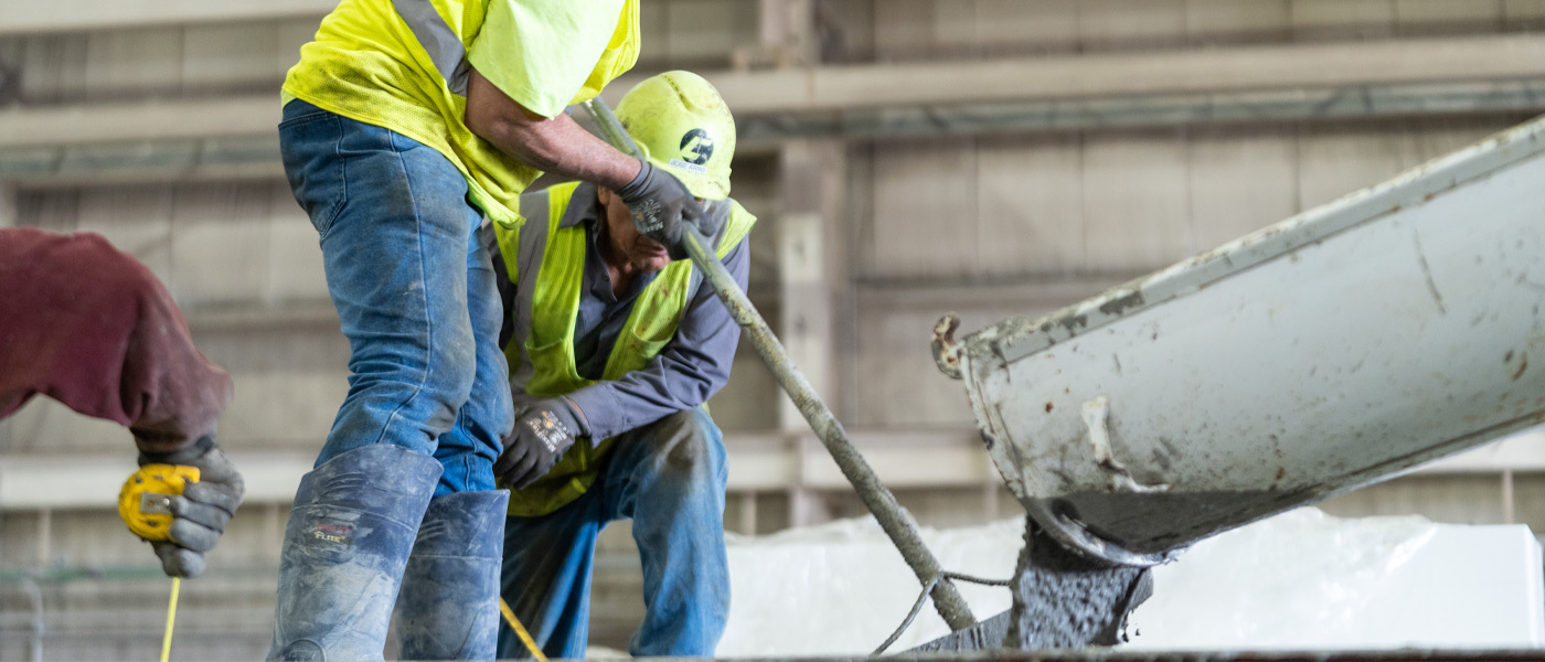 Two men wearing high-viz vests, hard hats, and blue jeans work with wet concrete in a warehouse.