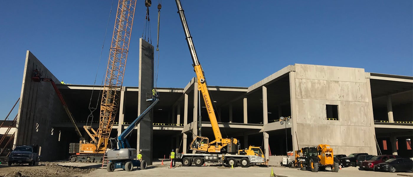 A crane raises a precast concrete exterior wall panel on the construction site of a parking garage for a multifamily residential building.